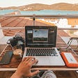Point of view shot of a young man’s hand on his laptop, with a portable charger, cup of coffee, DSLR camera, and smart phone on the table, around the laptop. He is overlooking the red tiled rooftops of nearby homes and the panoramic view of the ocean and a lush green hilly island ahead, with seagulls flying above on a clear sunny day with light blue skies