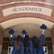 Three students in navy blue graduation gowns face a brick wall that reads “Academics,” while holding their caps aloft.