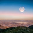 An out-of-this world view at the top of some mountain summit that is lushiously green but the vast view below is a valley full of brown ranges and craters while the sky is half dusk and half bright blue which the moon sits in it in ghost-like light — not fully lit and not fully erased.