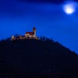 A castle on top of a hill, at night. There is a full moon in the sky, behind clouds.