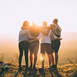 Four people wrap their hands around each other’s backs facing the sun in the mountains.