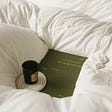 An olive green book of Shakespeare nestled in a white duvet with a cup and saucer