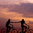 A man and woman holding hands as they ride bikes together at sunset
