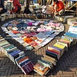 A lot of books stacks together on the ground to create a heart shape. There is one large heartshape on the outside and another smaller one inside of it. A couple people are looking at the books on the side of the heart.