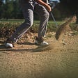 Male golfer executing a bunker shot-stance with weight on forward leg, iron passing through, and sand flying.
