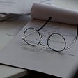 A pair of reading glasses sitting on top of a designer’s notepad on a desk with other papers.