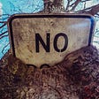 rectangular white “no” sign with black letters sticking out of a tree trunk