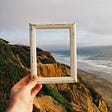 Person holding up empty frame to ocean view with cliff.