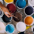 A person dipping a brush into a paint pot among many different colors.