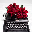Two blooming red flowers sit on top of a black typewriter