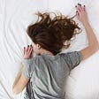 Woman in gray shirt laying on bed with white sheets wearing black nail polish