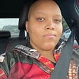 selfie of a light skinned Black woman in a car. She is looking at the camera with a blank expression. Her head is bald, and she’s wearing a hoodie that appears to have a red, brown, and gray pattern in the style of women’s armor.