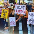 Photo of a protest about abortion rights. Signs read “Trust Women” and “Abortion is Health Care” and “Don’t Tread on Me.” One sign depicts a clothes hangar with the words “never again” inside.