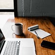 A Macbook laptop with a coffee mug, notebook and a phone sit on the table