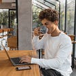 The photo of a short hair woman in a white long sleeve shirt drinking coffee while working on her laptop. ROMAN ODINTSOV/Pexels.