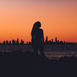 It’s sunset and there’s a silhouette of a woman overlooking the coastline of a city in the far distance.
