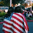 Two women wrapped in an American flag with their backs turned.
