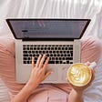 The picture shows a woman with a laptop on her knees, the woman is sitting on a bed. The picture is shot from above. We only see the woman’s legs, she wears soft pink pants. her left hand is on top of the laptop’s keyboard. In her right hand she is holding a cup of coffee.