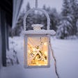 Winter scenery. In the background there is an open area covered in snow. Pine trees covered in snow in the farther back. In the foreground is a snow covered lantern, with a yellow glowing light inside.
