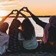 4 women facing the setting sun supporting one another with hands forming hearts above their heads.