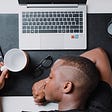 The One Motivator You Need Immediately to Stop Procrastinating. A person is seen sleeping on his desk. An empty coffee mug is in his left hand, and his laptop is open in front of him.