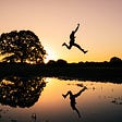 Silhouette of man jumping over water near a tree.
