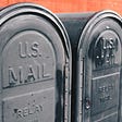 Two black U.S. mailboxes in front of an orange building.