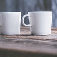 Two plain, white mugs, sitting on a wood surface with both their handles facing to the left.