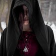 Woman looking all drama-ish in a woods with snow on the ground. She is wearing a cloak and a metal Celtic triquetra around her neck