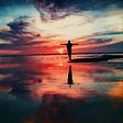 A man standing with outstretched arms in the shallows of the sea as the sun sets and cast colors across the water.