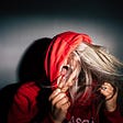 A woman, NOT Abigail Elphick, with a red hoodie pulled over her head and blond hair covering her face as she screams.