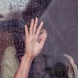 A woman with dark hair holds her hand up to a window with raindrops as if waiving goodbye.