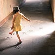 A little girl steeps through an outdoor corridor, her foot outstretched to reach the next paw print left imprinted on the ground.