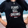 A woman stands with both hands at her hips in a superwoman pose. She is wearing a black T-Shirt with rolled-up sleeves. There is white writing on the front of the T-shirt that reads “Fears Kill Dreams. A red marker ink around the word dreams.