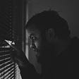 Man in a dim room peeking out through window blinds