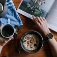 Person reading a book, with a cup of tea and a bowl of cereal in hands.