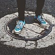 A person, wearing blue tennis shoes, standing on a manhole