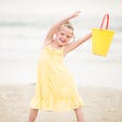 photo of young girl about six years old on the beach in a pale yellow dress holding a yellow plastic bucket in the air.