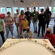 Photo of 12 attendees to the event standing and crouching behind a table displaying cupcakes arranged in the shape of an arc.