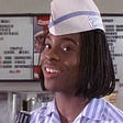 Kel Mitchell as Kel from “Good Burger” staring playfully into the camera in his light blue, striped, collared fast food uniform and paper cap.