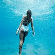 A Woman in a white bathing suit, as seen from behind. She swims towards the surface of the open water, her arms pointed down at at a 30 degree angle from her body. The water is light blue and their are bubbles within it.