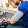 Woman using a blood pressure machine while having a telehealth visit. Photo by Henfaes/Getty Images