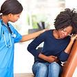 a Black female doctor with her hand on the shoulder of a young,Black woman who is grimacing with abdominal pain