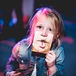 A young girl holding up a printed card that covers her mouth and fits right under her nose with the print of a puckered funny mouth as if she were being naughty and indirectly making faces at you.