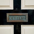 Close-up of a mail slot, with the word “LETTERS” engraved upon it, set within a black and white door.