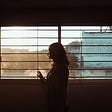 A woman looks out at buildings beyond a window, her body silhouetted against the light.