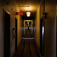 a dimly lit hallway that stretches far off into the istance. there is warm lighing that makes the walls look slightly yellow. doors are on either side of the hallway and continue down the entire stretch