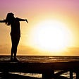 Silhouette of woman standing on a dock facing the sun with her arms outstretched at golden hour