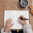 A moleskin notebook sits on a wooden table. A woman wearing rings and a long sleeve gray sweater is writing in the notebook with a black pen. Also on the table is a croissant with a bite taken out of it and a cup of black coffee in a brown coffee mug.