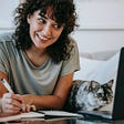 woman smiles at her laptop while taking notes. her cat, next to her, also looks at the laptop